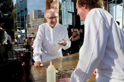 Alain Ducasse and another chef wearing chef's whites taste food and laugh