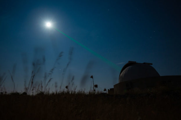 Night photograph of a silhouetted observatory sending a green laser to the moon