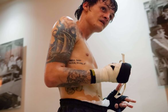 Shirtless, tattooed boxer, drenched in sweat, taking his wrist tapes off