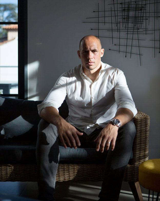 Portrait photograph of a man facing the camera, sitting on a wicker couch by a window