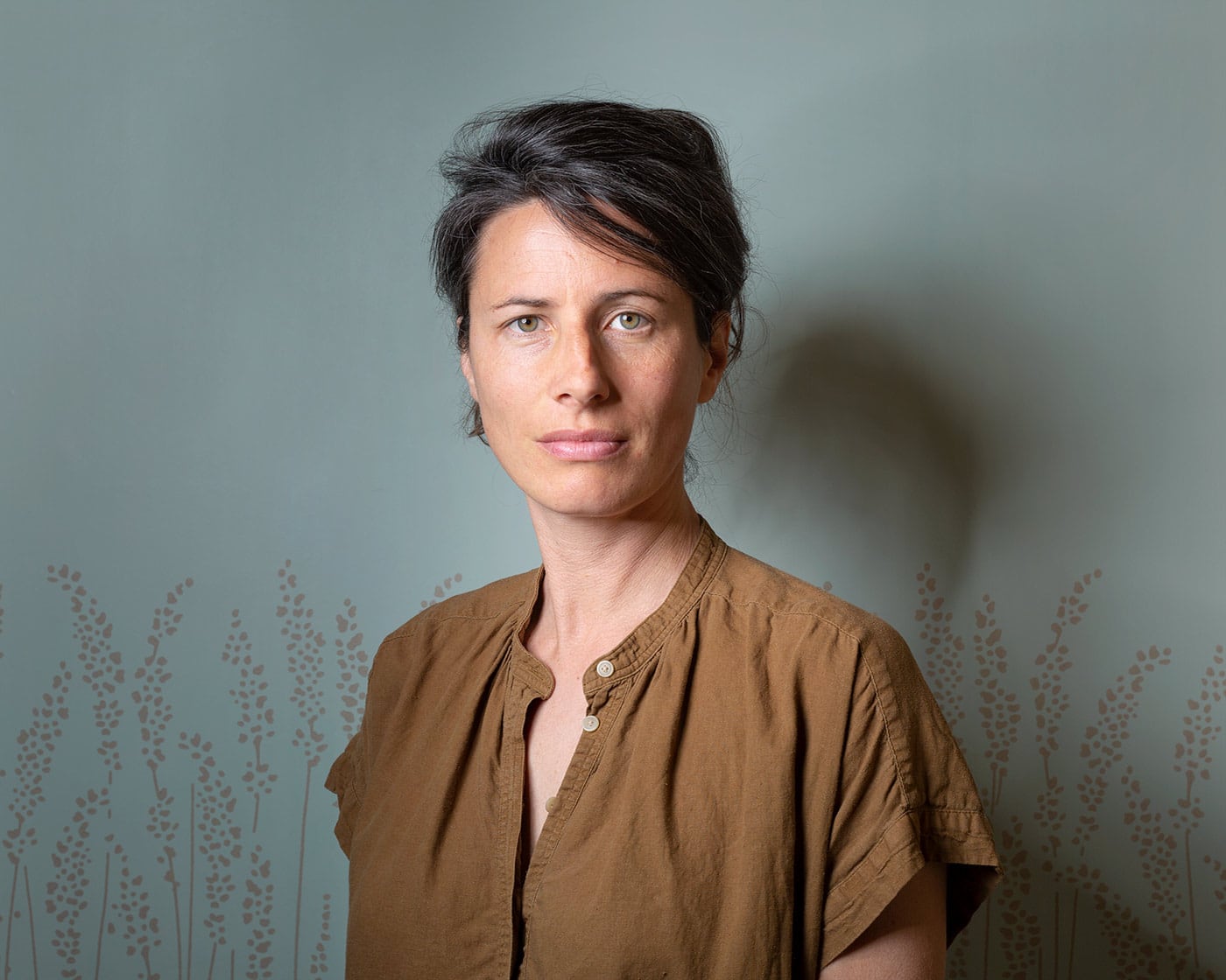Horizontal portrait of Rebecca Marshall in brown shirt in front of pale green-blue wallpaper with a grass pattern