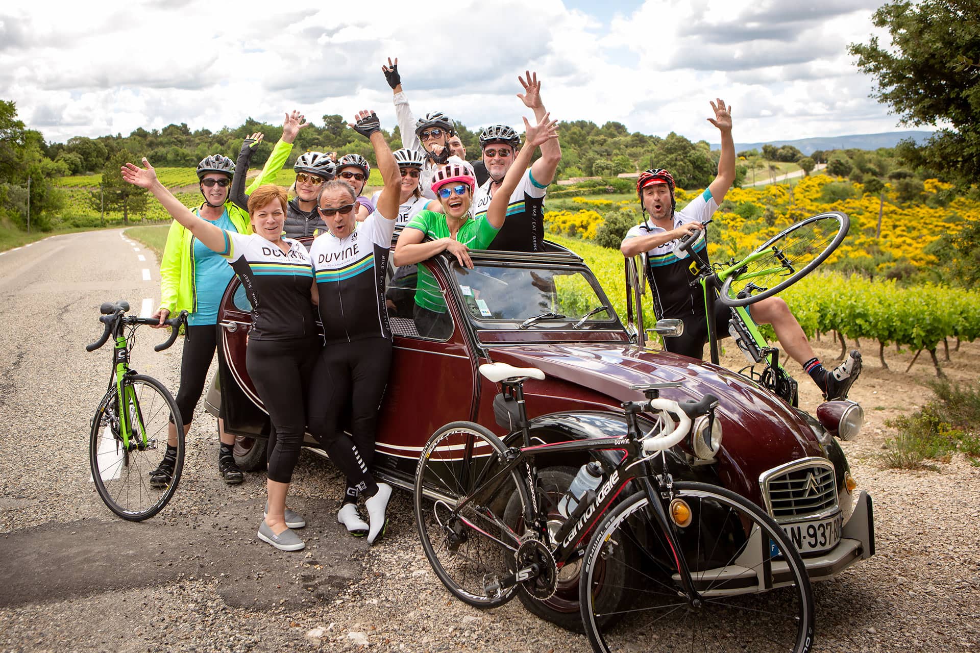 A group of cyclists in helmets gather around a Citroen 2 CV for a fun group photograph in the French countryside