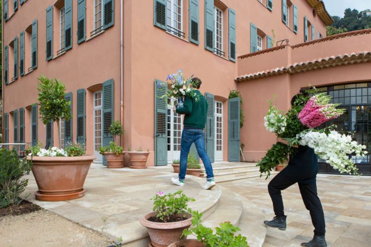 Two people carry armfuls of blossom up the steps of a large villa