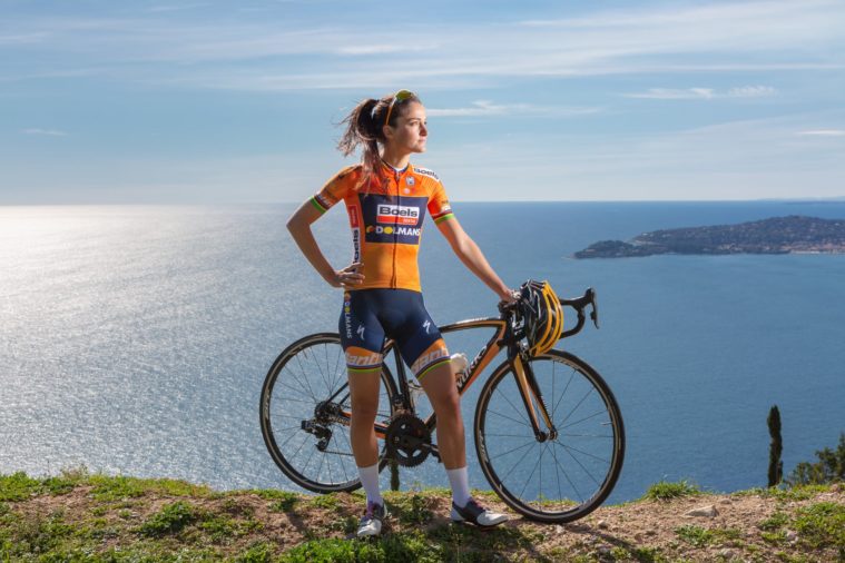Woman in cycling clothes stands at the top of a cliff above the sea with her bike