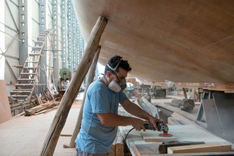 Man in a mask using a power tool to sand a piece of wood under the hull of a boat in a boatyard