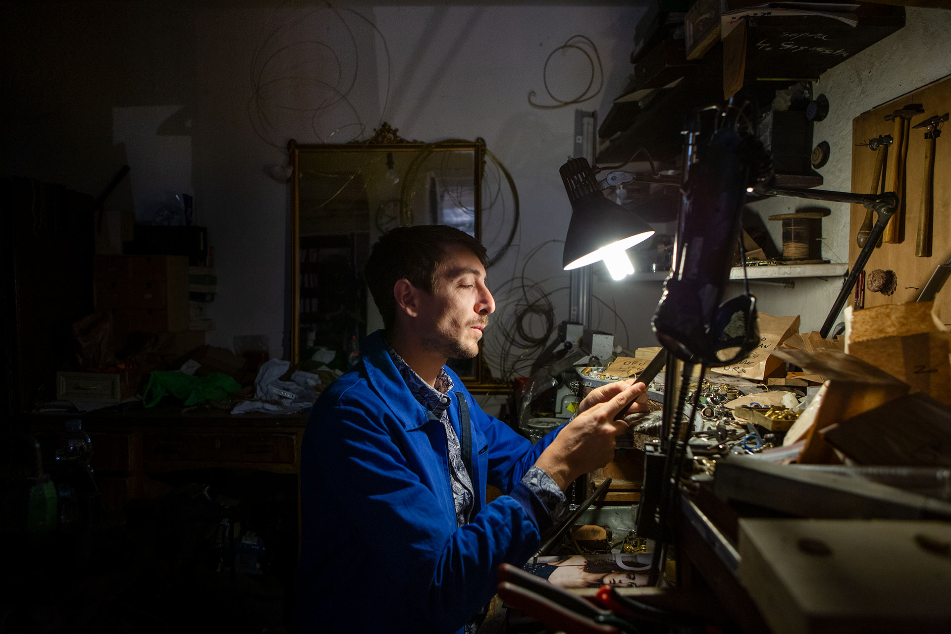 Man in a blue overall coat sits in a dark workshop under a lamp making jewellery