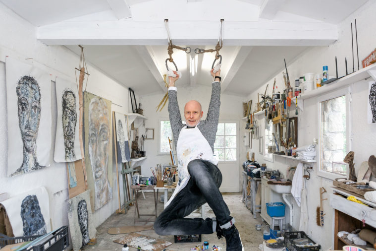 Environmental portrait of a man wearing a white apron covered in paint swinging from rings in the roof of a white-walled artist studio