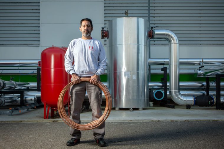 Man stands holding a coil of copper piping in front of a factory with pipes in the background