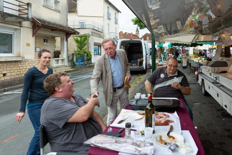 A man greets 3 people having lunch at a table behind a market stall