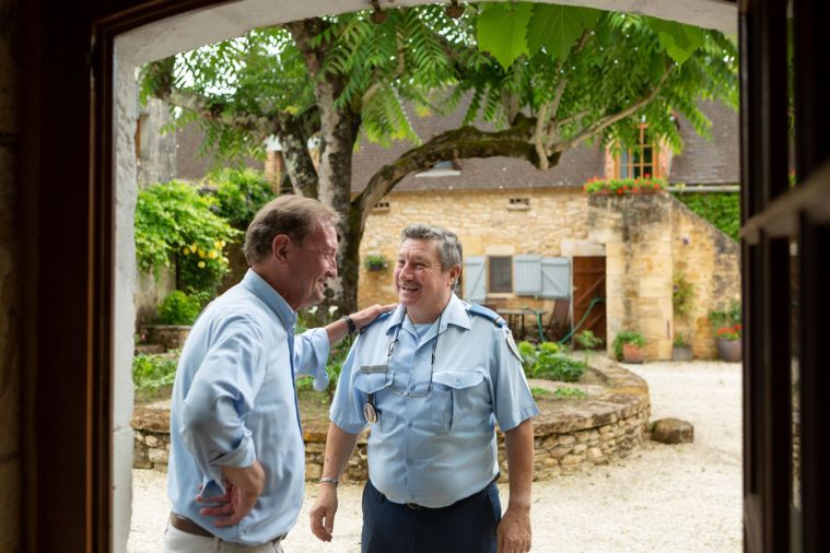 Two men, one a French policeman, greet each other in the doorway of a farmhouse