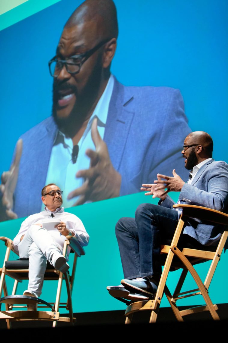 2 men, seated, speak on stage in front of a giant screen