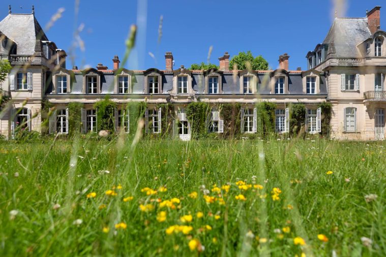 Photograph of a two-storey French chateau from the overgrown lawn