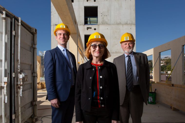 A woman and two men in yellow helmets smile to the camera on a building site