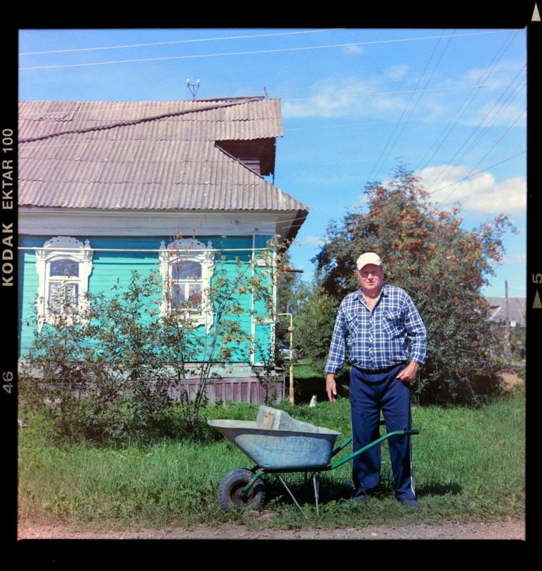 Photograph of a middle-aged man standing besided a wheelbarrow in the garden of his turquoise wooden house