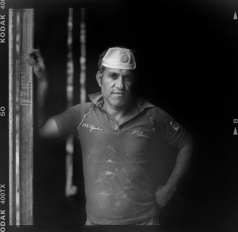 Black and white, medium format photograph showing a low key portrait of a workman, dust mask on his head