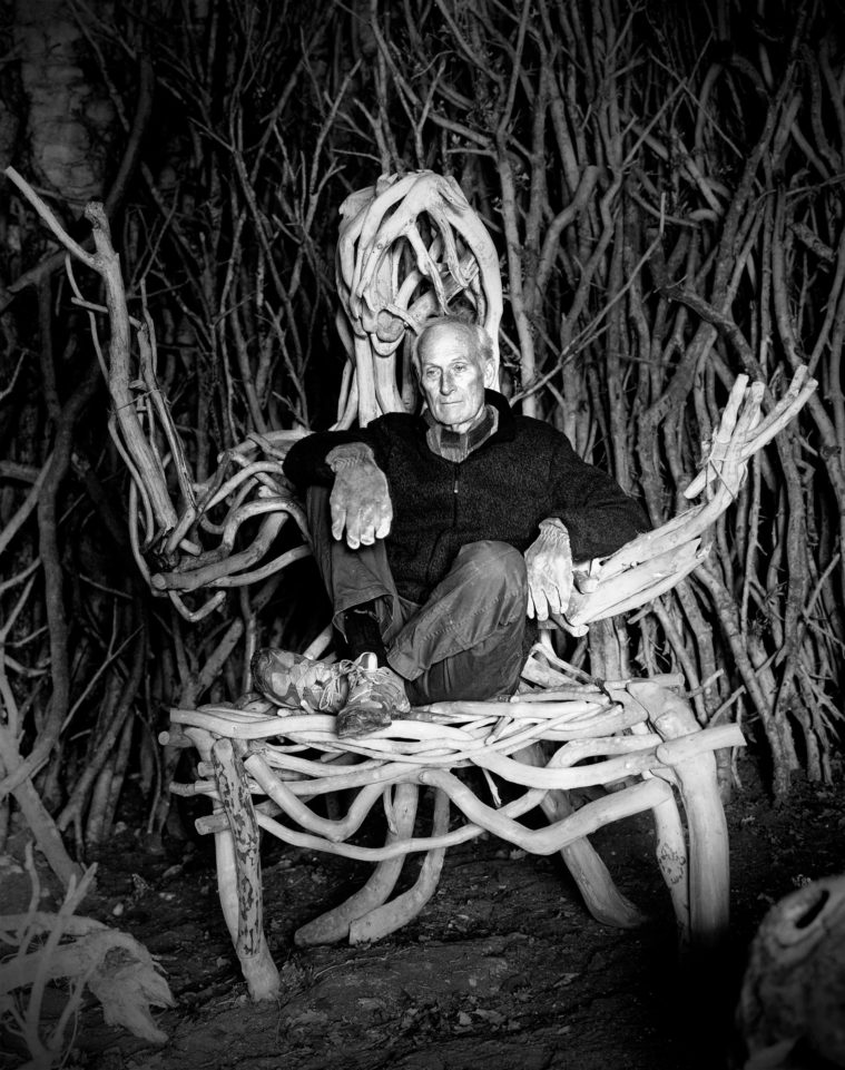 Black and white photograph of a man sitting in a large wooden chair shaped as a person in a woodshed