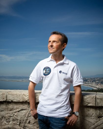 Man standing by a wall overlooking the coast wearing white T-shirt looks up at the blue sky