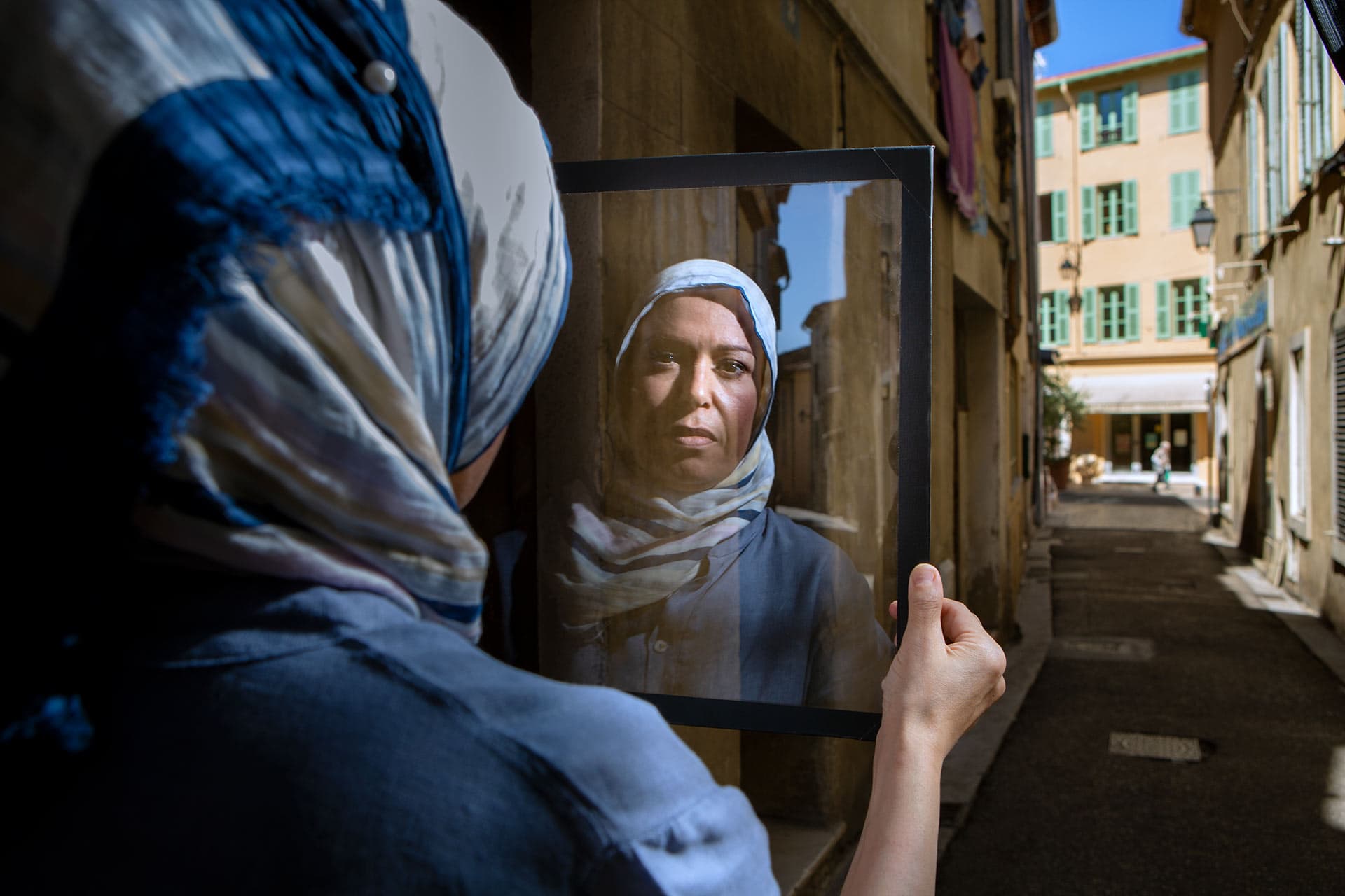 Woman in headscarf stands in a street and looks into her reflection in a sheet of glass