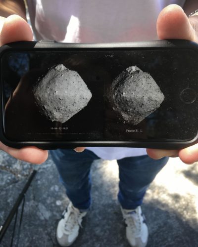 Asteroid Ryugu Close-up photograph of a person's hands holding a phone which displays two black and white photographs of an asteroid