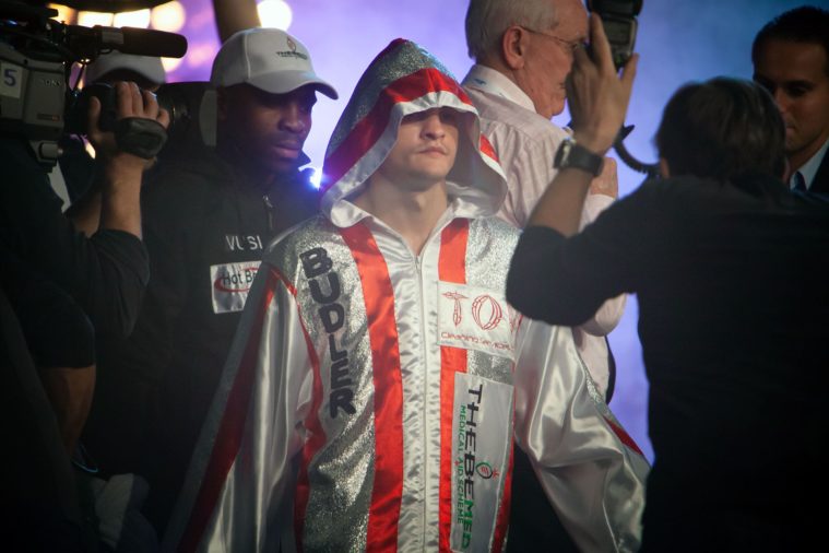 Hooded boxer arrives for a match surrounded by cameras and men
