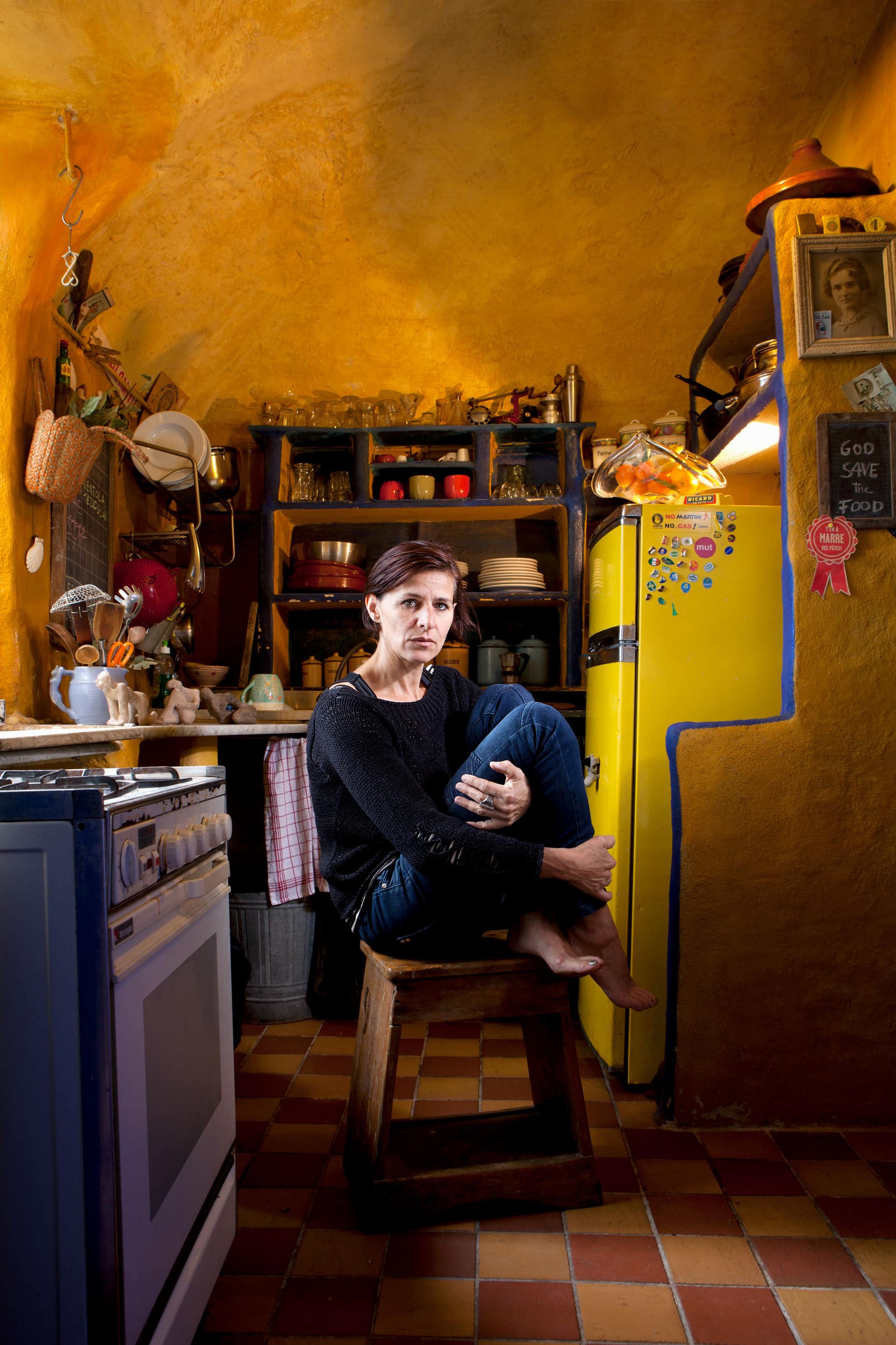 A woman hugs her knees sitting on a low wooden stool in a yellow kitchen