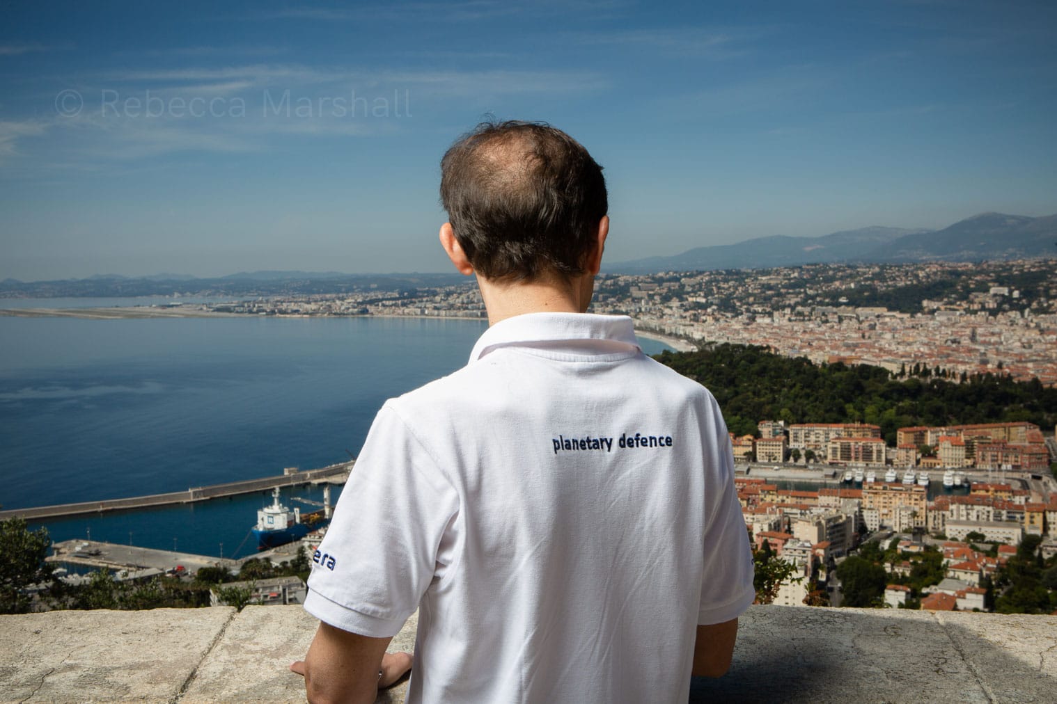 A man with his back to the camera, in a white T-shirt with 'planetary defence' printed on the back, looks down from a viewpoint over the coastal city of Nice