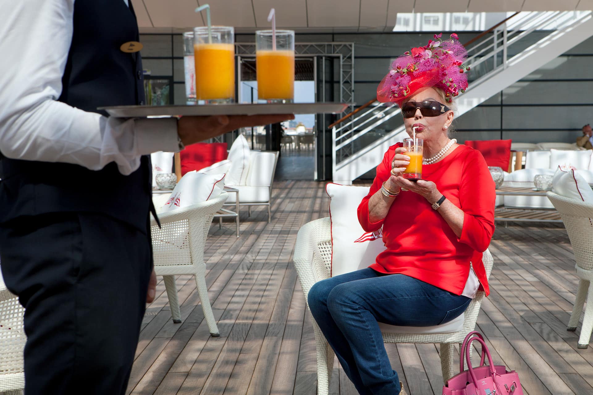 Woman wearing sunglasses, a red top and elaborate hat sips orange juice as a waiter stands in the foreground holding a tray