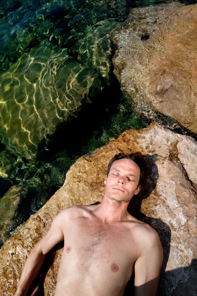 Man photographed from above lying on a roack beside the sea, seen naked from the waist up