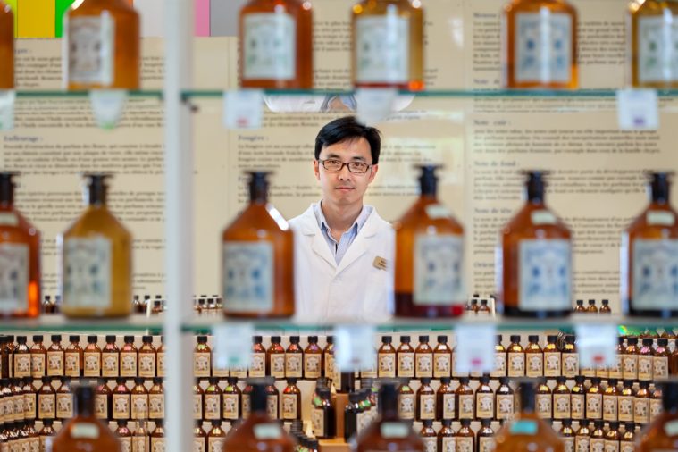 Portrait of a man wearing lab coat and glasses taken in between rows of out-of-focus brown perfume bottles