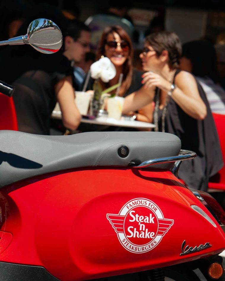 Close-up of a red Vespa scooter with Steak &#039;n Shake logo on it, 2 smiling women drink coffee in the background out of focus