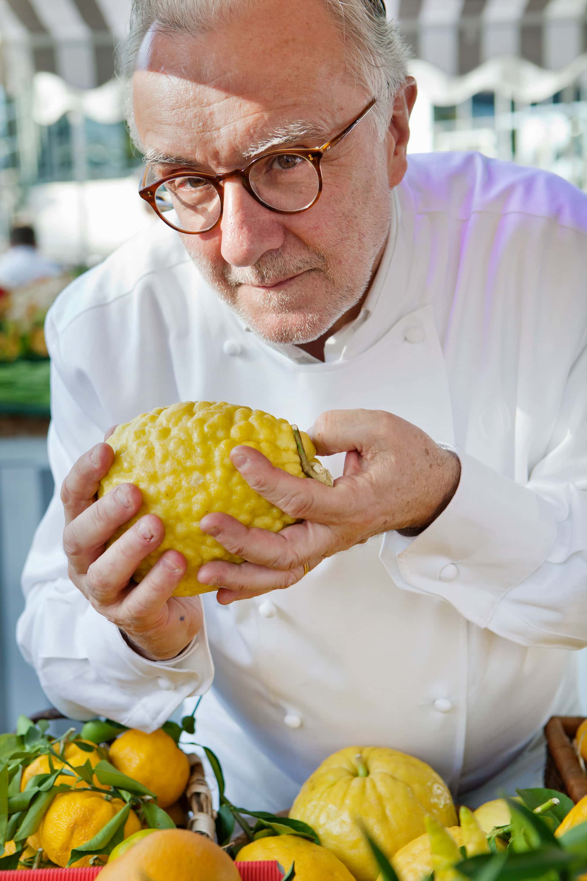 Close-up photograph of a chef holding a large yellow citrus fruit over a market stall display of lemons
