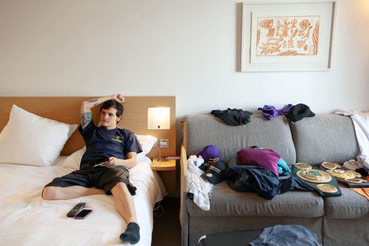 A young man lounges on a bed next to a sofa piled with clothes and boxing medal belts