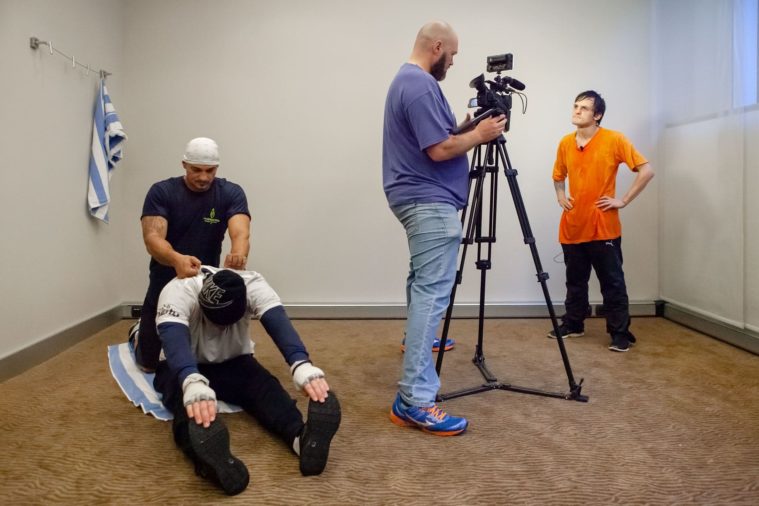 4 men in a room, two are stretching after exercise and the other very short man is being interviewed by a man with a video camera and tripod