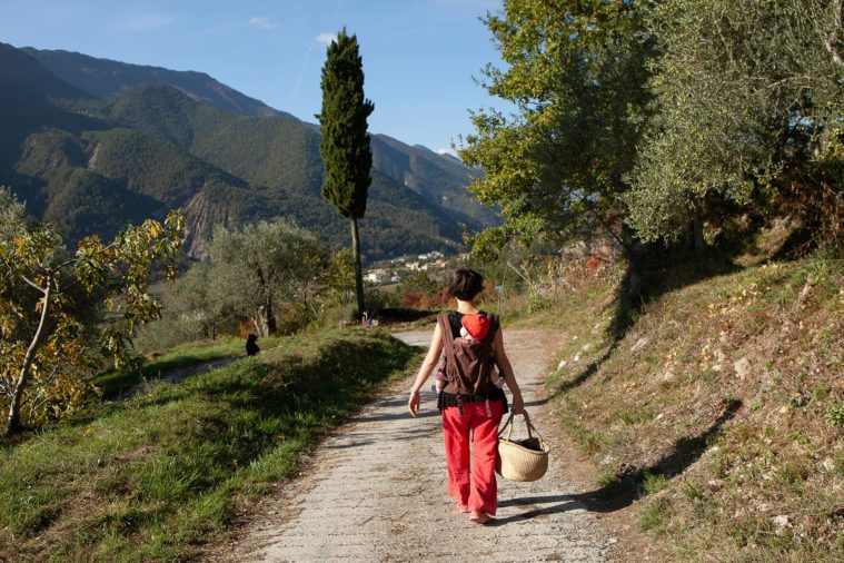 Woman with a baby strapped to her back walks along a country lane with a basket