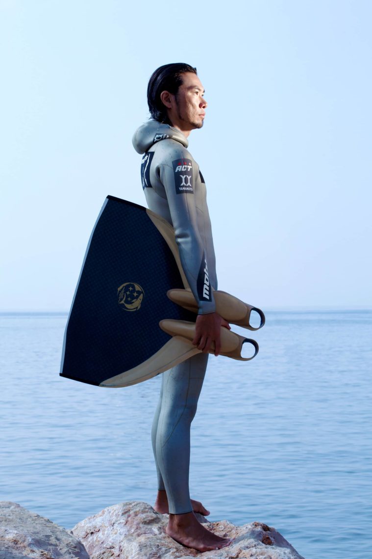 Man in a wetsuit in profile holding a monofin with the sea in the background