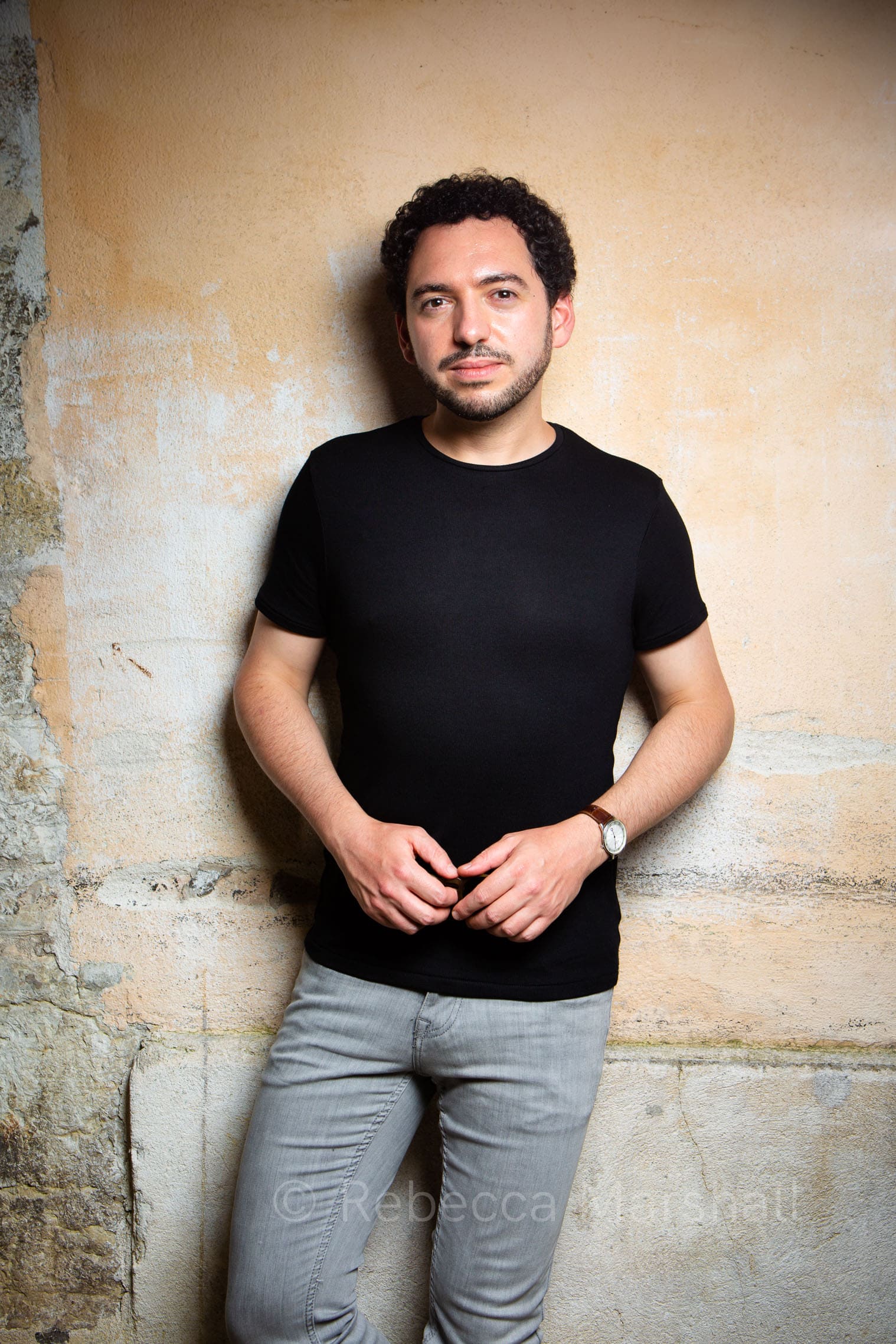 Dark-haired man in black T-shirt stands against a yellow wall facing the camera