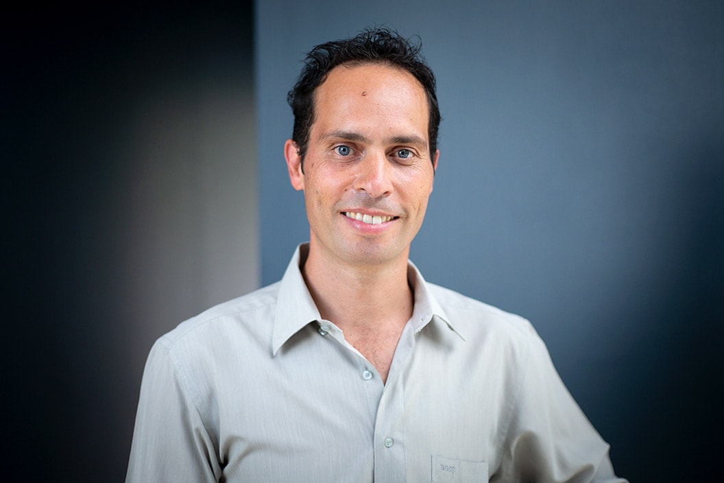 Portrait of a dark-haired man of European origin standing in front of a blue wall and smiling at the camera