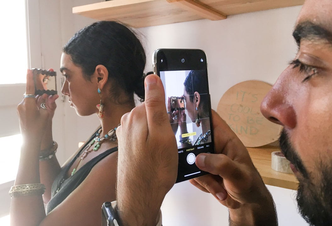 Close-up photograph of a man photographing a woman using a phone over her shoulder as she looks in a mirror