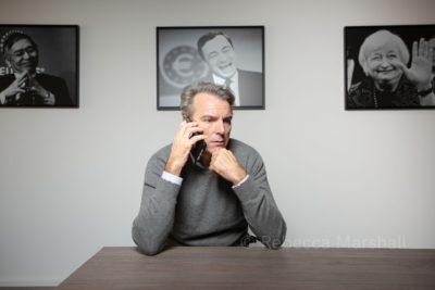 Photograph of a man on his mobile phone seated at a table, with 3 framed portraits on the wall behind him