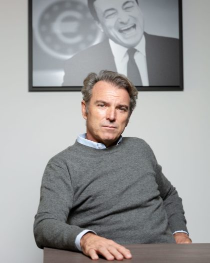 Portrait of a man in a shirt and grey jumper sitting in front of a framed picture of Mario Draghi on the wall behind