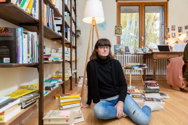 A woman in black jumper and jeans sits on the floor of a home office, surrounded by piles of books