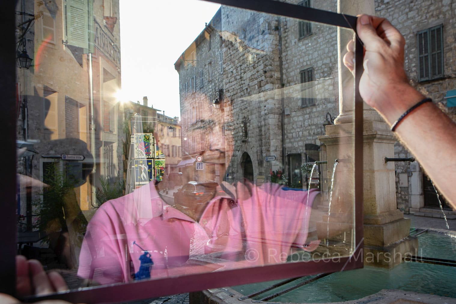 Photograph of the reflection of a man in a sheet of glass that he holds beside a fountain in a town square
