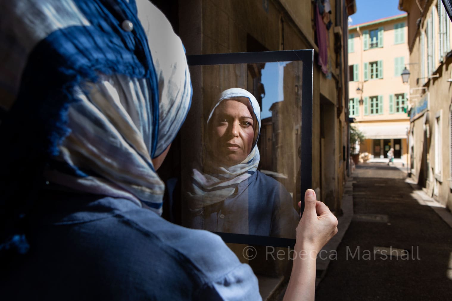 Photograph of the reflection of a headscarf-wearing woman in a sheet of glass that she holds in a quiet alley
