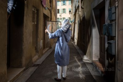 Photograph of the reflection of a headscarf-wearing woman in a sheet of glass that she holds in a quiet alley