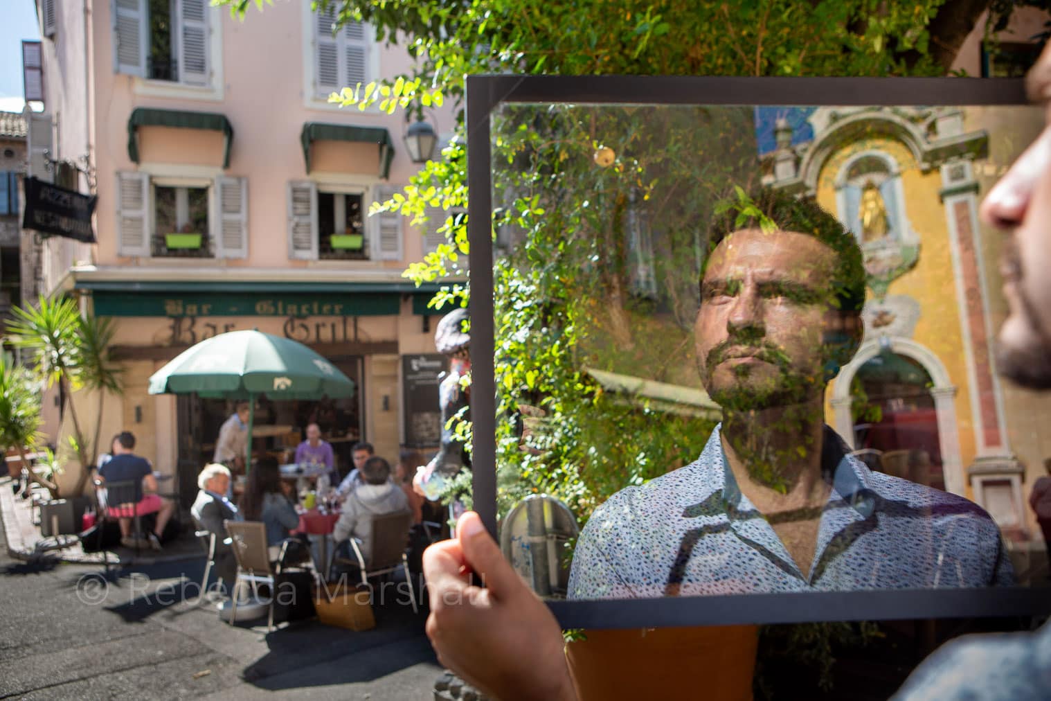 Photograph of the reflection of a man in a sheet of glass that he holds in a busy town square