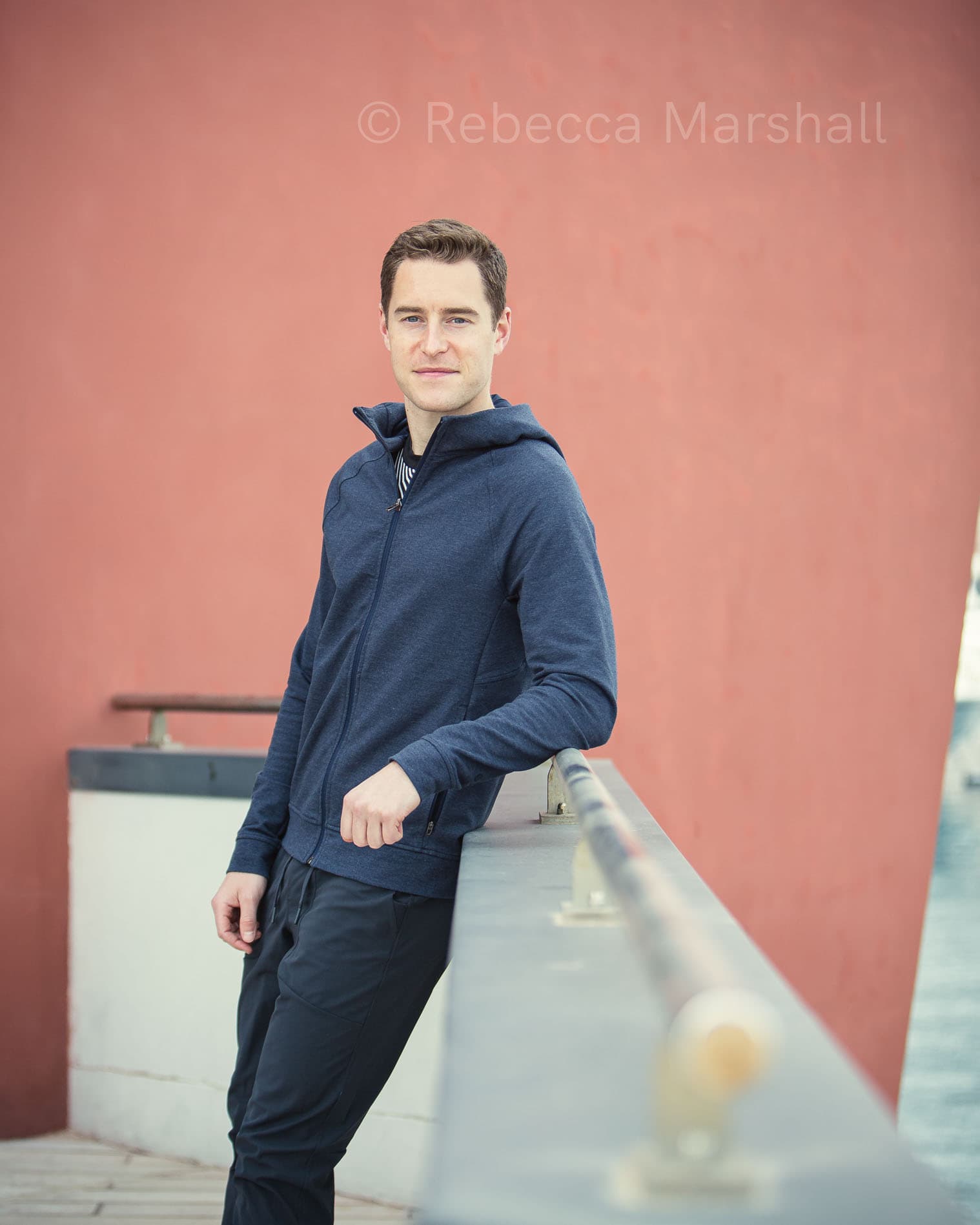 Portrait of a man in a blue tracksuit leaning on a railing in front of a red wall