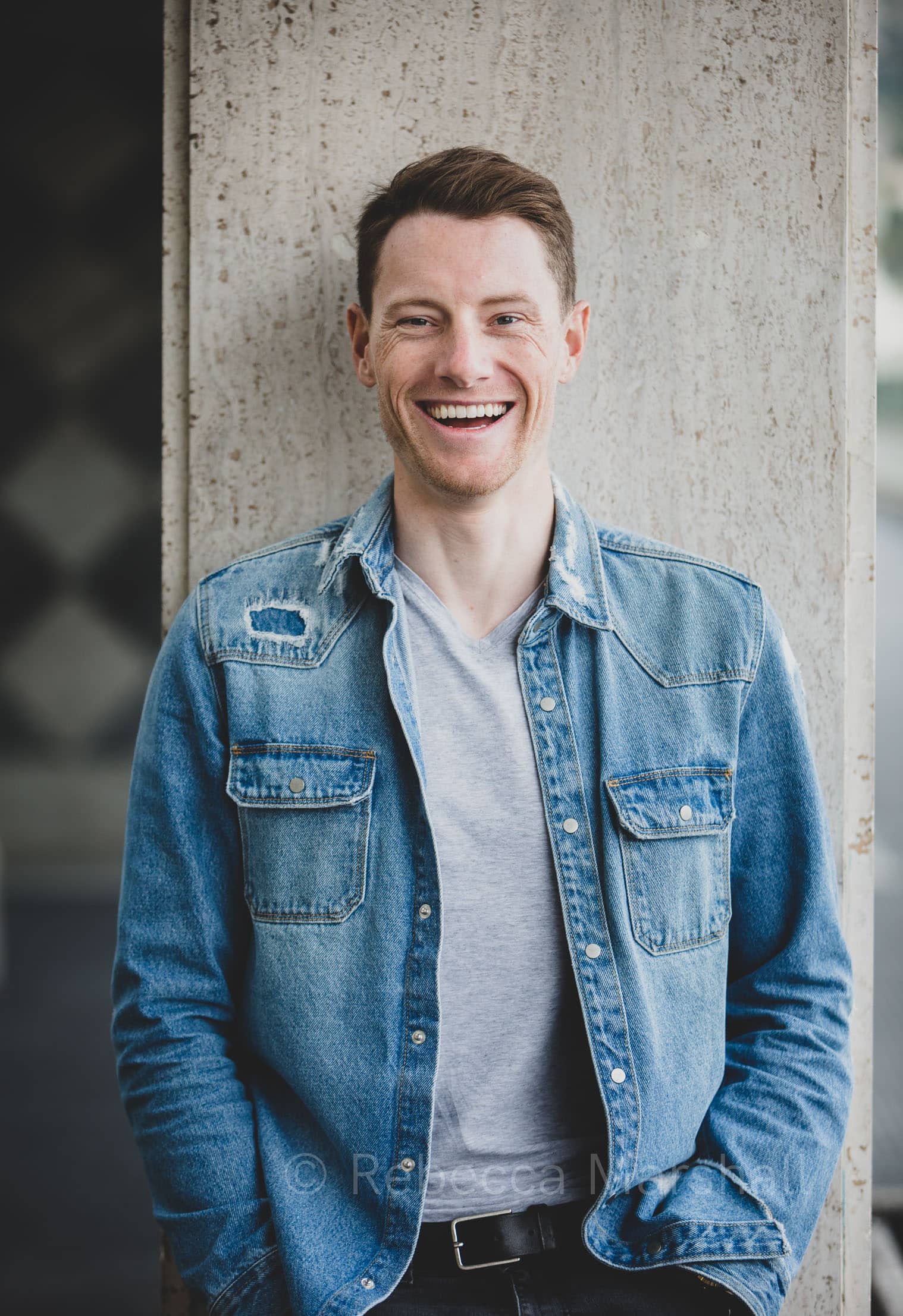 Photograph of Sam Bennett wearing a blue denim jacket, leaning against a wall and laughing