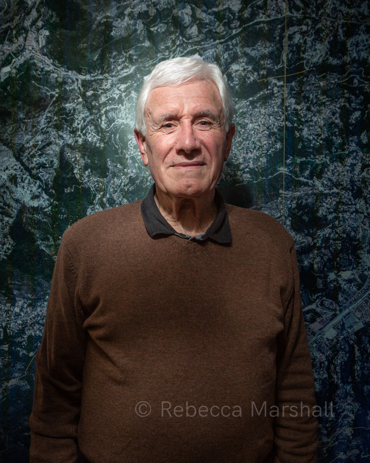 Portrait of a white-haired man standing before a satellite photo