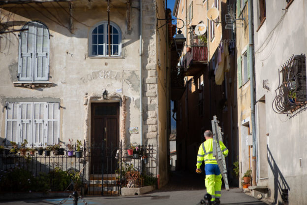 Photograph of a man in fluorescent yellow workwear carrying a ladder along a village alley