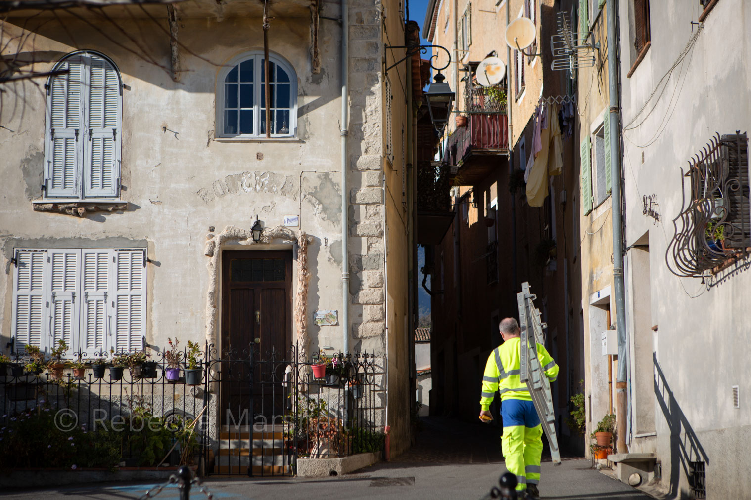 Photograph of a man in fluorescent yellow workwear carrying a ladder along a village alley